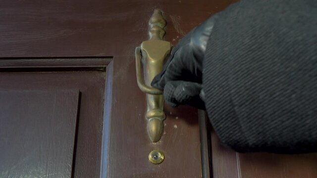 Closeup POV shot of a person wearing a black glove and coat, using a hand to strike the brass door knocker on an old wooden front door, four times. 