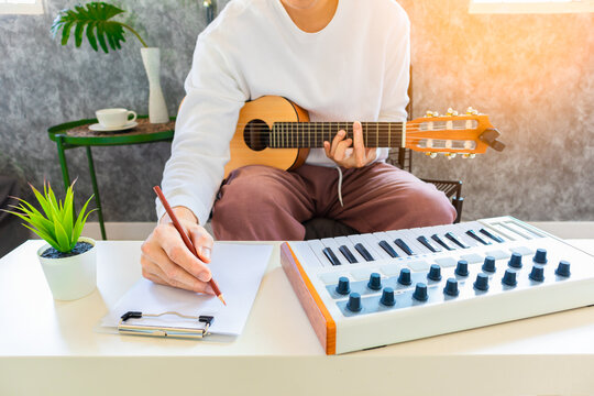 male amateur songwriter playing acoustic guitar and writing a song on white paper in living room