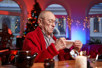 Bald and serious elder man sitting at table and holding a letter in cosy and decorated room with christmas tree.