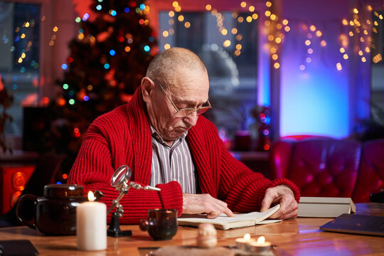 An Old Man Weared With Eyeglasses Sits At Table Writing Some Letter And Xmas Cards In Cosy Room Decorated With Lights And Christmas Tree