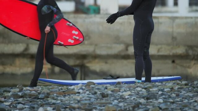 Russian winter surfing. Female surfer runs with red surfboard behind another surfer warming up on the coast of the Black sea in Russia