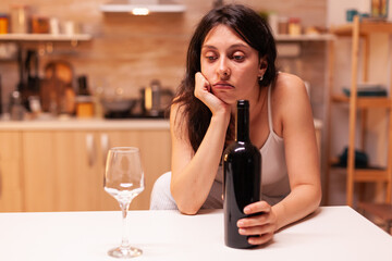 Woman sitting alone with a bottle of red wine in kitchen because of edpression. Unhappy person suffering of migraine, depression, disease and anxiety feeling exhausted.