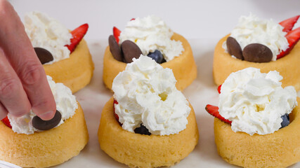 Biscuit cakes with fresh berries, chocolate, and whipped heavy cream. Woman preparing dessert, mini biscuit cups with strawberries and blueberries,  close up on white background
