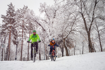 Father and son ride bicycles in the winter park. Bearded man and boy on bicycles spend family weekend in beautiful snowy winter park