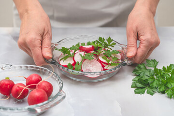 Fresh sliced radish with fresh green parsley close up on glass plate on light grey background. Woman serving vegetable salad