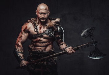 Fototapeta na wymiar Muscular and violent viking barbarian with hairless head poses in dark background holding his two handed double axe.