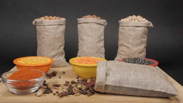Gunny bag/jute sack full of dried whole spices falling on a wooden table with lentils. Collection of various colorful raw dal/pulses sack kept on wooden table against a black background