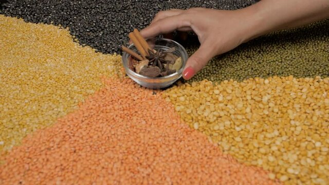 Woman's hand keeping a transparent bowl with Indian whole spices on various lentils. Closeup shot of a set of different types of lentils on a table with a small bowl of spices - nutritious protein-...
