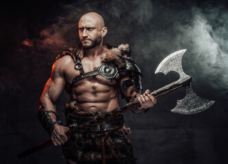 Fototapeta na wymiar Dressed in antique light armour with fur bald and violent viking with muscular build poses in dark atmospheric background holding two handed double axe