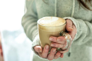 A woman in a warm sweater is holding in her hands a clear glass cup with hot aromatic coffee in her hands. Concept of a cozy morning. Closeup
