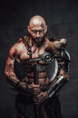 Portrait of a nordic bald warrior armed with two handed axe and in dark armour with fur in atmospheric dark background looking at camera