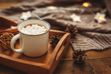 A cup of hot drink of chocolate or cocao with marshmallows on a wooden table in a cozy interior with a plaid. Cozy winter concept