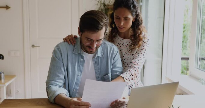 Smiling young family couple reading paper correspondence letter, feeling satisfied with pleasant news, discussing taxes refunds, utility bills or financial bank loan mortgage payments, planning budget