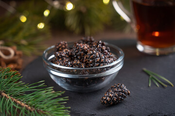 Fototapeta na wymiar Siberian vegan dessert of dried pine cones canned in syrup served in bowl with cup of tea on dark wooden background with pine branches and garland lights at winter holidays at kitchen. Horizontal