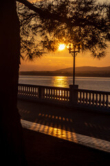 Summer Sunset in Golden tones. Silhouette of a balustrade. Shadow of the balustrade on the embankment. Back lighting. The sun sets in the mountains and is reflected in the sea. Pine branch in the