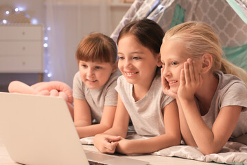 Fototapeta na wymiar Cute little girls lying in wigwam and watching cartoons on laptop