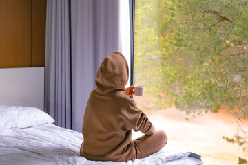 Rear view of a woman wearing a hoody drinking coffee looking at forest view through the window