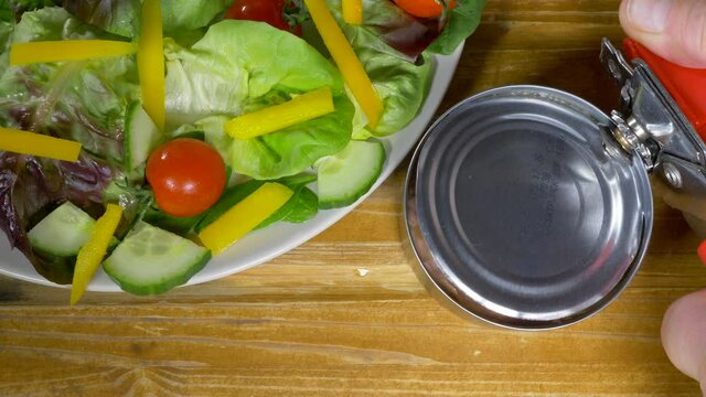 Closeup POV overhead shot of a man&rsquo;s hands using a can opener on the lid of a tin of chopped skipjack tuna in water, next to a plate of salad, on a pine worktop / counter.