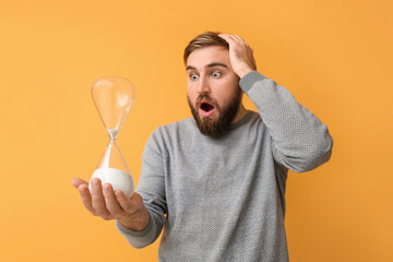 Shocked young man with hourglass on color background