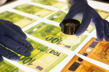 Hands in rubber gloves holding magnifying glass over banknotes closeup