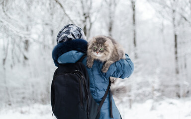 Woman carrying a cat on her shoulder outside, in the winter forest. Fluffy cat with a sad expression on his face, looking away