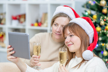 Happy mother and her daughter wearing Christmas hats having video call with tablet computer and toasting with Champagne while celebrating Christmas at home