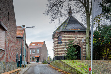 Fototapeta na wymiar Empty street between houses and the Chapel of Mary in de Nood on a small hill with its gray dome, a Christmas tree next to the door, cold and cloudy day in Stein, South Limburg, the Netherlands