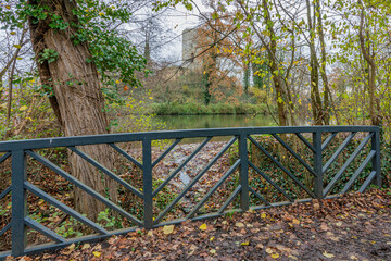 Footbridge over a stream that flows into a lake with Stein Castle in the background, autumn trees, leaves on the ground and climbing plants, cloudy day in Stein, South Limburg, Netherlands