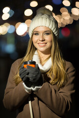 a blonde girl, in the evening, against the background of the lights of the fair with a drink in an orange glass, a white hat and a beige coat walks on the street. smile