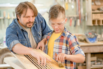 Father teaches his young son how to paint a wooden board with a paintbrush in a carpentry workshop
