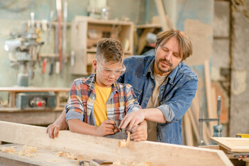 Father teaches his young son to plan wood in a carpentry workshop