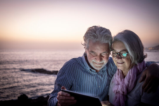 A senior couple white-haired looking at digital tablet in the dusk sitting on the beach with horizon over water on background. Happy retirement concept together