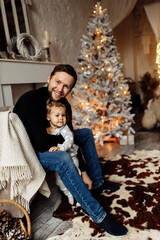 Caring parent with little daughter spend winter holidays at home. Loving father sitting at the chair near Christmas tree, hugging cute toddler, enjoy happy moments, New Year celebration concept