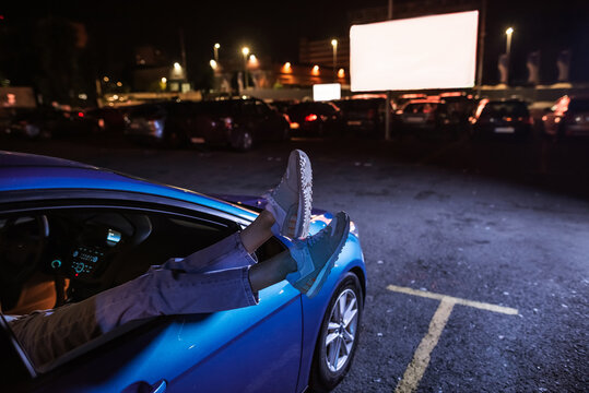 Come here for fun. Close up of woman s legs out of the car window parked in front of a big white screen at drive in cinema