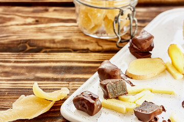 Candied ginger slices in dark chocolate on a white plate on wooden table. Dehydrated oriental fruits. Healthy sweets. Antioxidants.