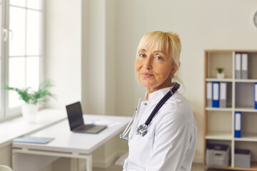 Close up portrait of senior tired female doctor looking at camera while standing in medical office.
