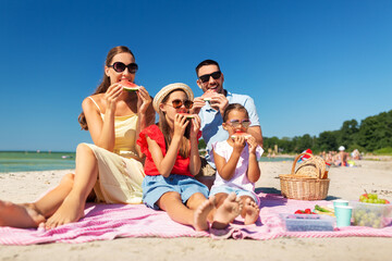 family, leisure and people concept - happy mother, father and two daughters having picnic on summer beach and eating watermelon