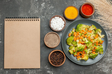 Healthy meal with brocoli and carrots on a black plate and spices next to notebook on gray table
