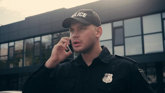 Policeman in uniform and cap talking on smartphone on urban street