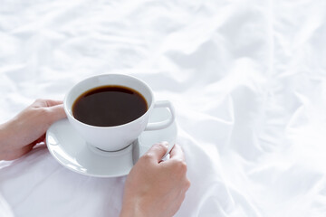 white coffee cup hold by woman hand on bedsheet in weekend with sun light. concept wake up in morning with warm drink to awake self in vacation or holiday relax time in bedroom with copy space.
