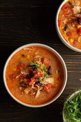 Spicy mexican soup with beans and corn, tomatoes, chicken breast and bean sprouts, bowl with soup and napkin on a dark wooden background, top view