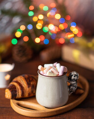  ceramic cup with cocoa and marshmallows and a baked croissant on a brown table