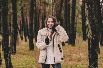 Beautiful young woman model appearance with long curly hair holding a Christmas candy in the form of a heart and smiling while being in a pine forest