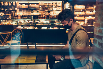 Young man sitting in coffee shop at store front in the city center in the evening, wearing the face mask to avoid virus infection and to prevent the spread of disease in time of coronavirus