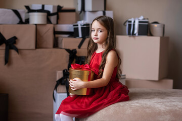 A girl in a red dress is holding a gift. Against the background of a heap of other gifts. Christmas photo