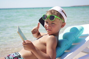 Tanned child shows a thumbs up resting on the beach and playing on a tablet while lying in a sun lounger. Cute boy uses tablet on the beach on a Sunny summer day.