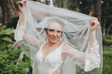 A smiling, sweet bride holds a veil with her hands and looks out from under it. Wedding close-up portrait, photography.