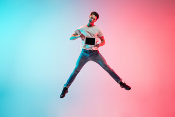 Naklejka na ściany i meble Tablet screen. Young caucasian man's jumping on gradient blue-pink studio background in neon light. Concept of youth, human emotions, facial expression, sales, ad. Full length, copyspace.