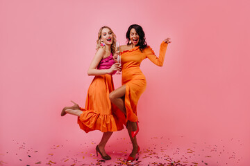 Two attractive girls dancing at party. Indoor full length photo of carefree female models posing on pink background.