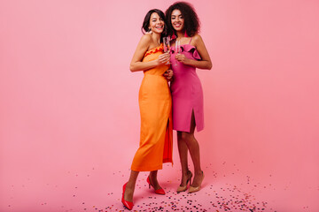 Full-length portrait of two gorgeous girls in party dresses. African female model drinking champagne in studio.